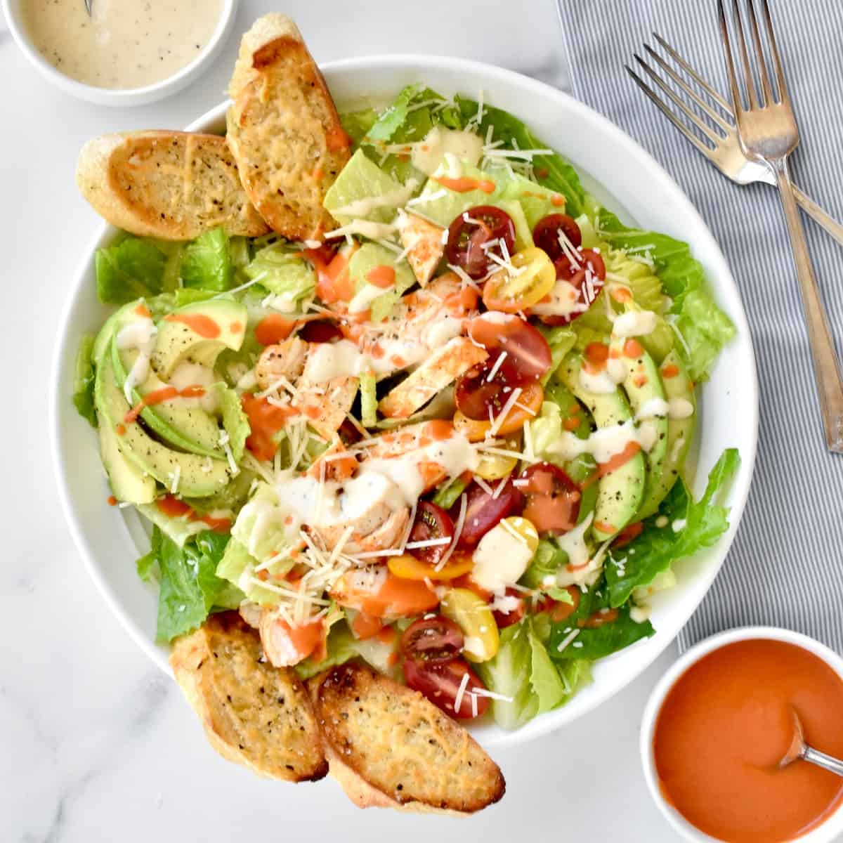 A white bowl with buffalo chicken caesar salad, with two forks, a dish of caesar dressing, and a dish of buffalo sauce to the side.