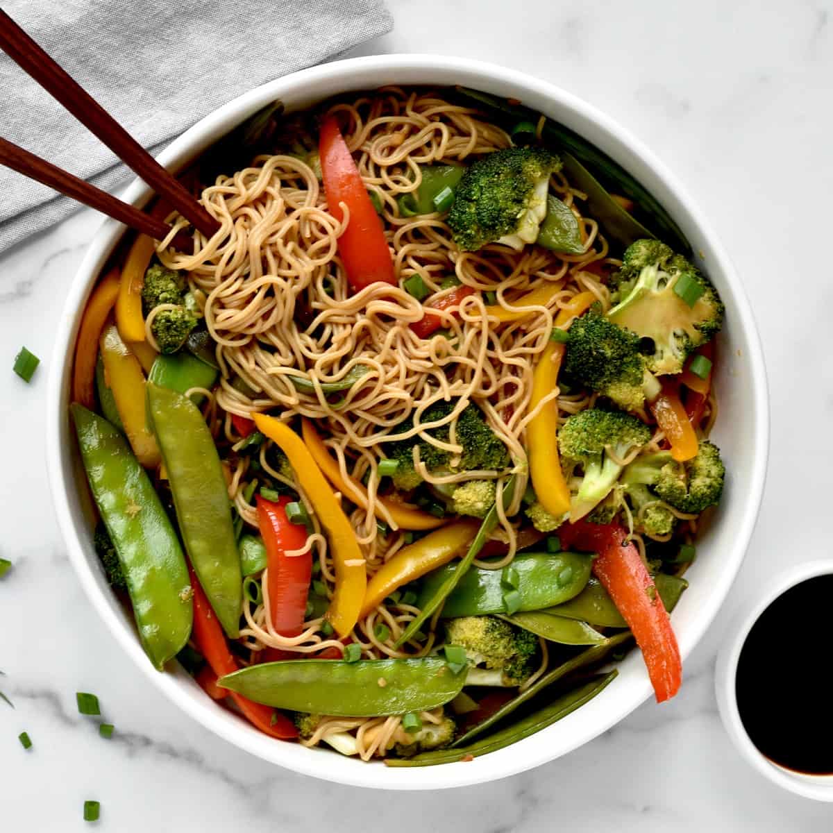 A bowl of ramen noodles with vegetables in stir fry sauce, with a side of soy sauce and two chopsticks twirled around some noodles.