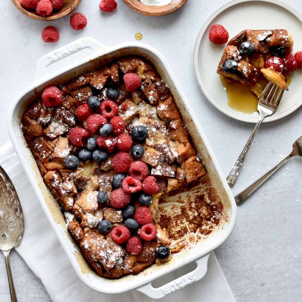 A white casserole dish with french toast casserole sprinkled with powdered sugar and fresh berries. A plate next to the dish has one serving of french toast casserole with maple syrup, and two forks and a serving spoon are to the side.