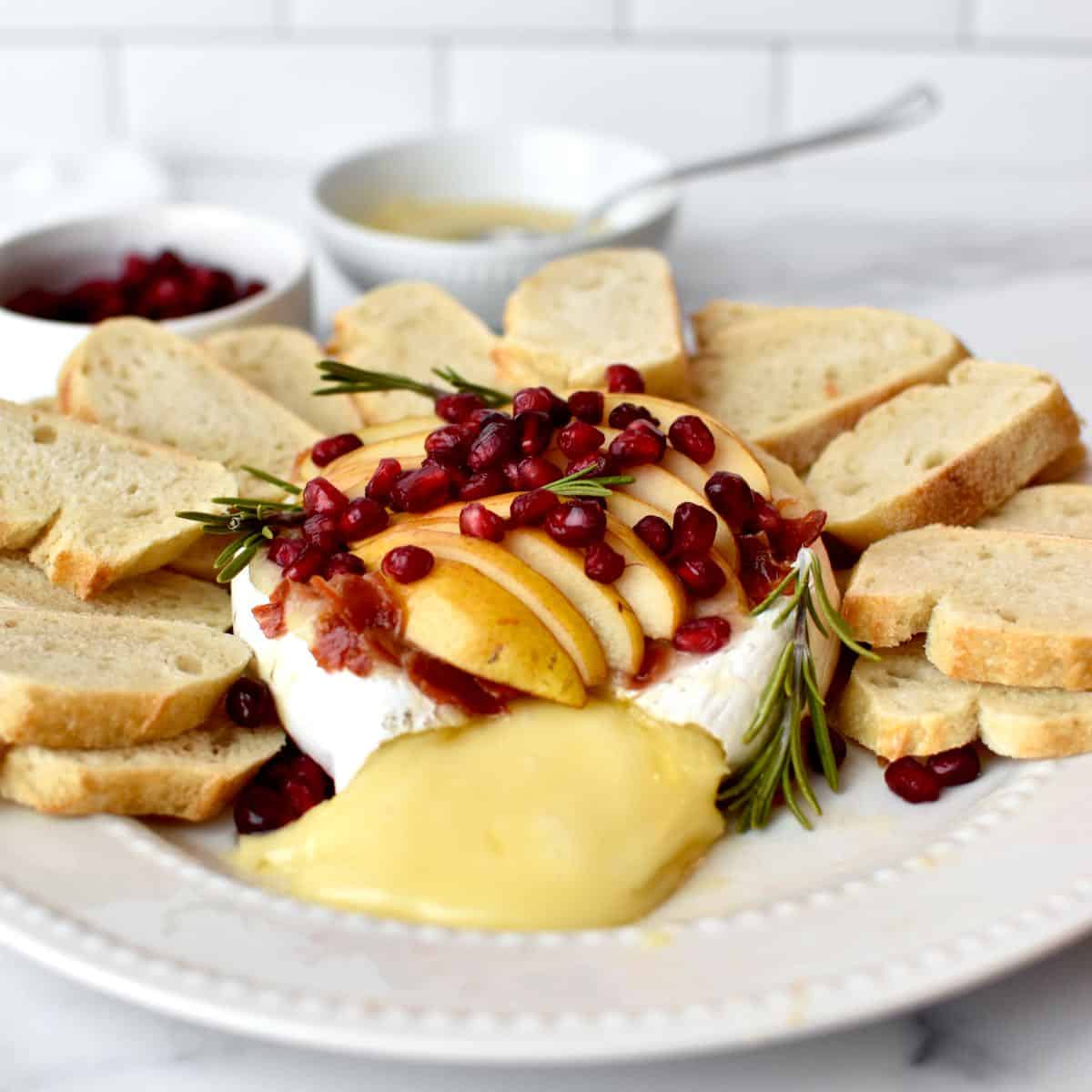 Side view of baked brie, topped with pears, prosciutto and pomegranate. The brie is surrounded by sliced bread and is oozing melted cheese.
