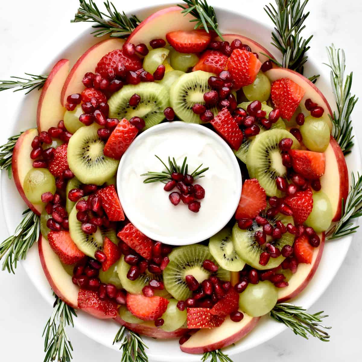 A fruit salad arranged in a wreath shape on a white plate, with a bowl of yogurt sauce in the middle.