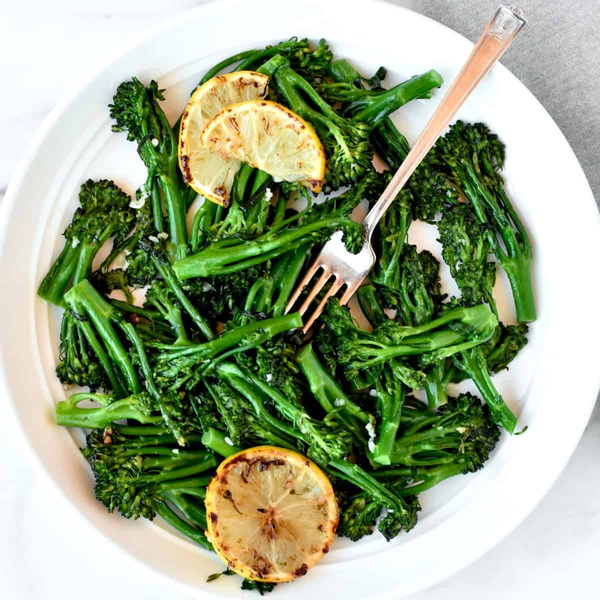 A white bowl with sauteed broccoletti, with two lemons arranged in the bowl and a fork sticking out of it.
