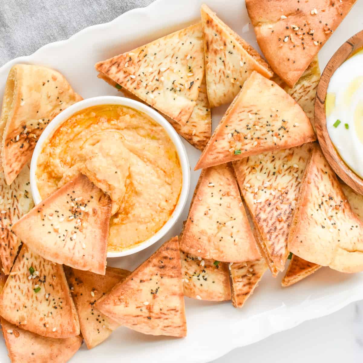 A close up of air fryer pita chips on a white tray. The tray contains the chips and a bowl of hummus with one pita chip dipping into the hummus.