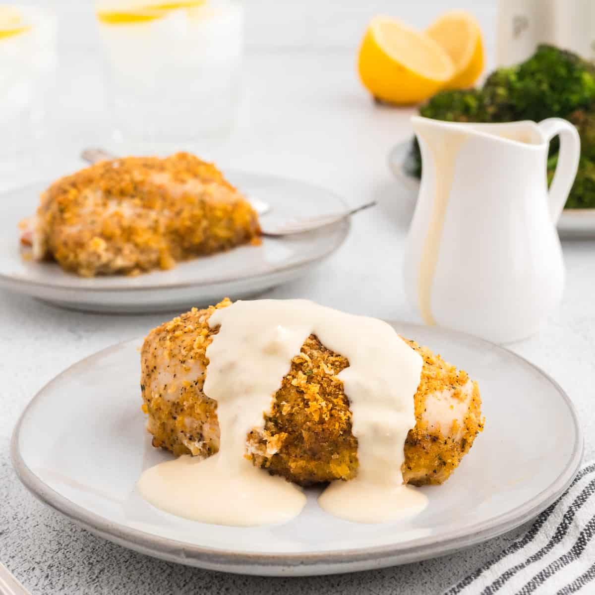 A breast of chicken cordon bleu on a grey plate, drizzled with creamy dijon sauce. Another plate of chicken and a plate of broccoli are in the background.