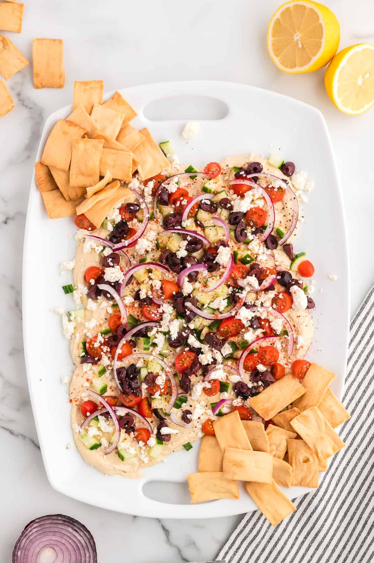 A white platter on a marble background with loaded hummus topped with cucumbers, cherry tomatoes, red onion, feta cheese and kalamata olives. Pita chips are arranged on the sides of the platter.